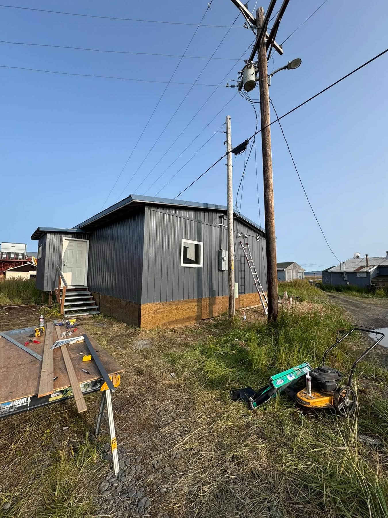 Gray metal-sided home on a sunny day, with a ladder against the wall, utility poles and a work table of tools in the foreground